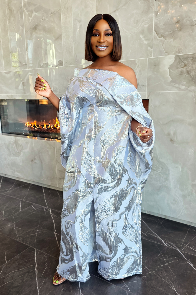 A woman wearing a light blue and silver abstract-patterned off-the-shoulder maxi dress, standing in front of a modern fireplace with a marble-tile wall and floor.