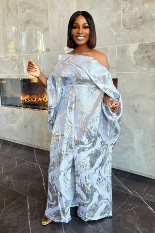 A woman wearing a light blue and silver abstract-patterned off-the-shoulder maxi dress, standing in front of a modern fireplace with a marble-tile wall and floor.