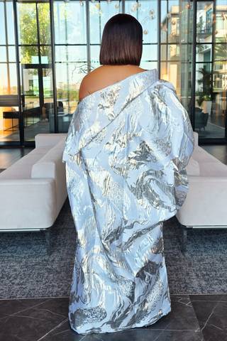 A woman viewed from behind wearing a voluminous off-the-shoulder wrap or robe with a metallic silver and white abstract marbled pattern, standing in a modern lobby with white sofas and large glass windows.