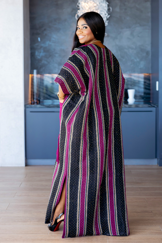 A woman wearing a long, flowing kaftan dress with vertical stripes in black, magenta, and beige, photographed in a modern interior setting.