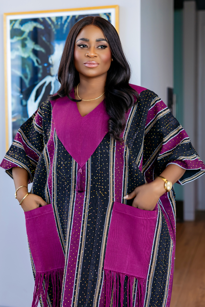 A woman wearing a long kaftan dress with vertical black, pink, and beige stripes, featuring a solid magenta V-neckline, large magenta patch pockets with fringe, and matching fringe details on the sleeves, standing in a modern interior setting.