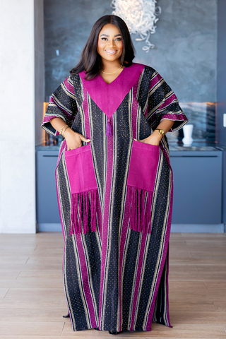 A woman wearing a long kaftan dress with vertical black, pink, and beige stripes, featuring a solid magenta V-neckline, large magenta patch pockets with fringe, and matching fringe details on the sleeves, standing in a modern interior setting.