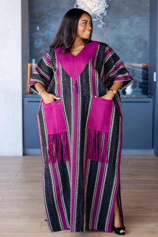 A woman wearing a long, loose kaftan dress with vertical stripes in black, white, and magenta, featuring a solid magenta V-neckline and matching pocket flaps with fringe details, standing in an indoor setting.