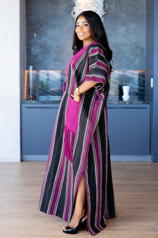 A woman modeling a long, flowing kaftan dress with vertical stripes in black, magenta, and white, paired with a solid magenta inner dress and black heels, set against a modern interior background.