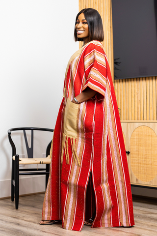 A woman wearing a red and beige striped kaftan dress with a tan inner top, standing in a modern room with a wooden chair and slatted wood wall paneling.