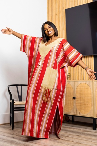 A woman modeling a red and beige striped kaftan dress with a gold patterned shawl draped over her shoulders, posing in a modern living room setting.