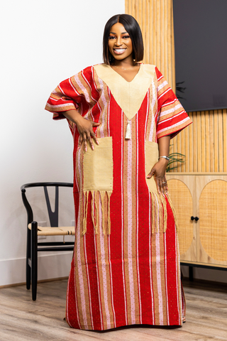 A woman modeling a long red kaftan dress with vertical beige and white stripes, featuring a beige V-neckline, large front patch pockets, and fringe details, set against an indoor background with a wooden chair and cabinet.