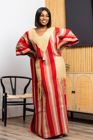 A woman modeling a long kaftan dress with vertical red, beige, and white stripes, standing in a modern room with a chair and wooden cabinet in the background.