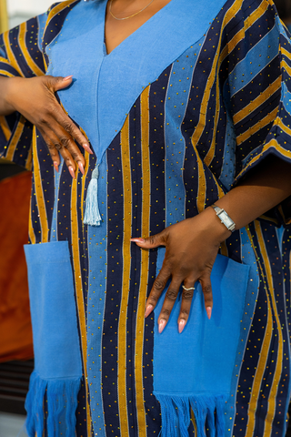 A close-up of a person wearing a blue and navy striped kaftan dress with gold dotted vertical stripes, solid blue V-neck panels, and matching blue fringe pockets.