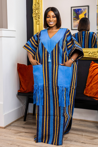 A woman modeling a blue, black, and gold striped kaftan dress with solid blue pockets and a tassel tie, standing in a room with a mirror and orange pillows.