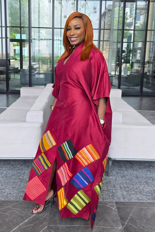 A woman wearing a vibrant magenta satin kaftan dress with colorful African wax print (Ankara) panels on the hem, standing in a modern lobby with white sofas and glass windows.