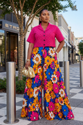 Woman in a pink top and colorful floral pants standing on a sidewalk with a store in the background.