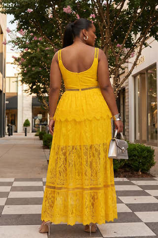 Woman in a yellow lace dress walking on a sidewalk with trees and buildings in the background.