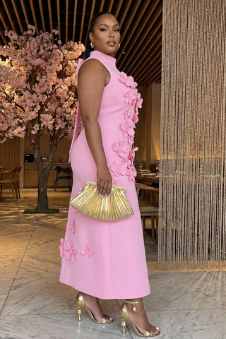 Woman in a pink dress with floral details holding a gold clutch in an indoor setting.