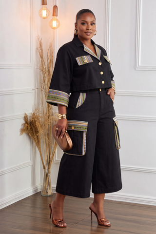 A woman models a black two-piece jumpsuit set with colorful patterned trim details, standing in a decorated room with wood floors and wall paneling.
