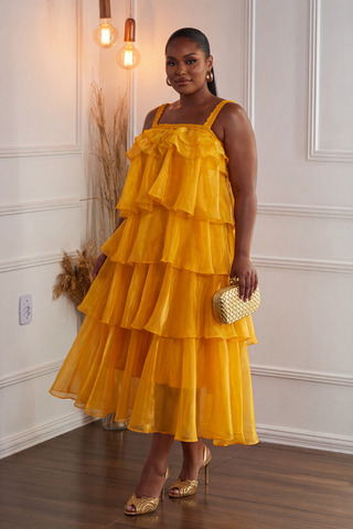 A woman models a bright yellow tiered ruffle dress with spaghetti straps, paired with gold metallic heels and a small gold woven clutch, standing in a white-paneled room with warm hanging lights.