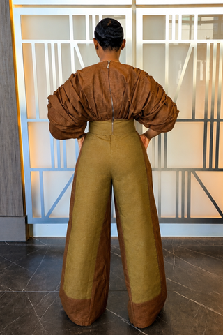A person viewed from behind wearing a brown and olive green wide-leg jumpsuit in front of a modern geometric glass wall.